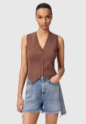 Kvinde iført brun ærmeløs, ribbet vest med knapper og slidte denimshorts med kæde ved lommen, stående foran ensfarvet baggrund.