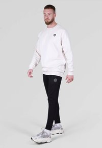 Világos bézs pulóver fekete logóval, amelyet fekete melegítőnadrággal párosítanak, szintén egyező logóval. A modellen szürke sportcipő és fehér zoknik vannak.