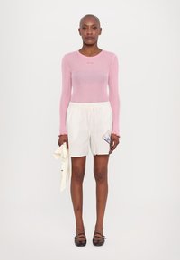 Lys rosa ribbet langermet topp kombinert med hvite stripete shortser med en sidepute. Modellen holder et kremfarget stofftilbehør.