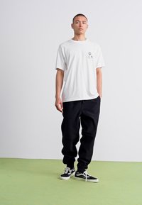 Tricou alb din bumbac cu guler rotund și trei imprimeuri grafice mici, asortat cu pantaloni de trening negri, largi și teniși negri.
