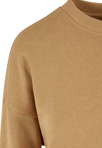 Hellbraunes Sweatshirt mit geripptem Kragen, weicher Textur und lockerer Passform mit überschnittenen Schultern. Keine sichtbaren Muster oder Hardware-Details.