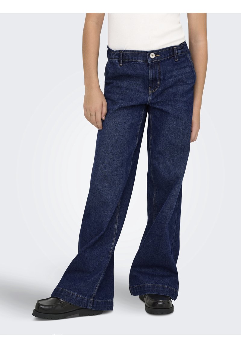 Personne portant un jean large bleu foncé, des chaussures noires épaisses et un haut blanc sur un fond uni clair.