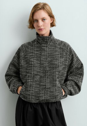 Jeune personne aux cheveux blonds courts porte une veste zippée texturée noire et blanche, les mains dans les poches, contre un mur gris.