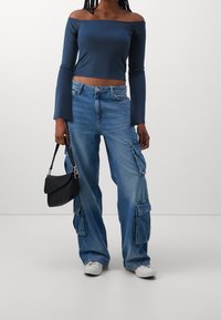 Kvinna som bär en marinblå off-shoulder långärmad topp, baggy blå cargojeans, vita sneakers och håller en liten svart handväska vid sin sida.