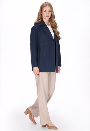 Laivastonsininen kaksirivinen takki suurilla napeilla, yhdistettynä beigenvärisiin leveälahkeisiin housuihin ja beigeihin korkokenkiin, valkoista taustaa vasten.