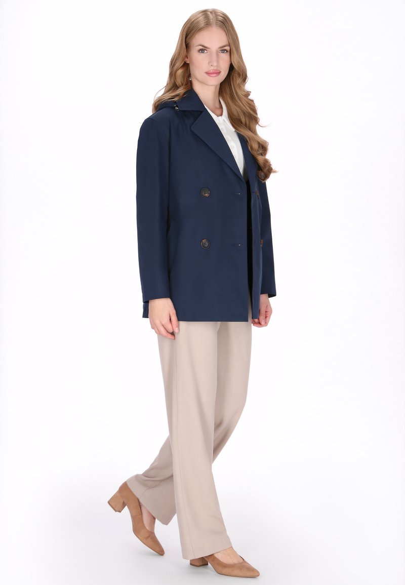 Námořnický kabát se dvěma řadami knoflíků a velkými knoflíky, kombinovaný s béžovými širokými kalhotami a béžovými podpatky, na jednobarevném bílém pozadí.