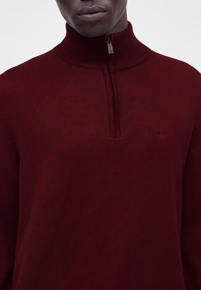 Homme portant un pull en maille côtelée couleur bordeaux avec un col montant zippé et un logo discret sur la poitrine gauche.