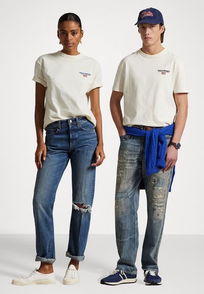 Dva modela nose casual odjeću: krem majicu, plave traperice s ispucalim detaljima i tenisice. Muški model nosi mornarsku kapu i plavi pulover vezan oko struka.