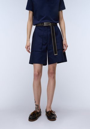 Person iført marineblå shorts, et matchende marineblåt bælte med synligt mærke, sorte snørebåndssko og en marineblå top; tatovering synlig på venstre ben.