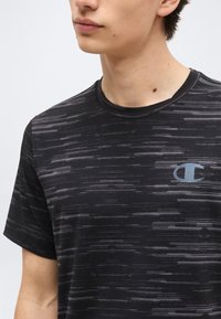 Νεαρό άτομο που φοράει ένα μαύρο και γκρι T-shirt με μοτίβο και στρογγυλή λαιμόκοψη, με μικρό φωτεινό μπλε λογότυπο Champion στο αριστερό στήθος.