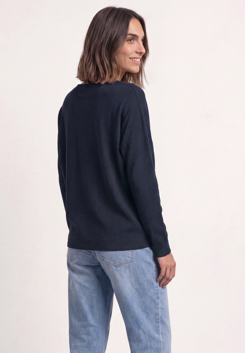 Femme aux cheveux mi-longs souriante, portant un haut bleu marine à manches longues et un jean bleu clair, debout devant un fond uni.