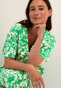 Cvečni potiskani kombinezon v zeleni in beli, z kratkimi rokavi in globokim V-izrezom. Mehak, lahek material z sproščenim krojem.