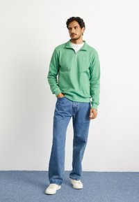 Pullover verde texturizado com meia abertura, apresentando um bolso frontal. Combinado com jeans de perna reta azul claros e ténis brancos.