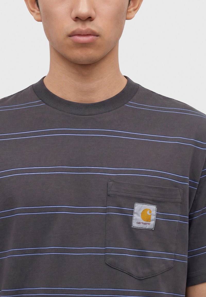 Giovane che indossa una t-shirt a righe grigio scuro con una tasca sul petto che presenta una toppa con il logo giallo Carhartt.