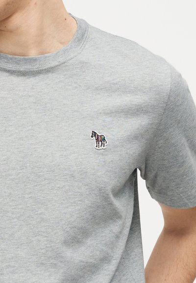 Κοντινό πλάνο ατόμου που φοράει ανοιχτό γκρι μπλουζάκι με μικρό πολύχρωμο έμβλημα ζέβρας στο αριστερό στήθος.