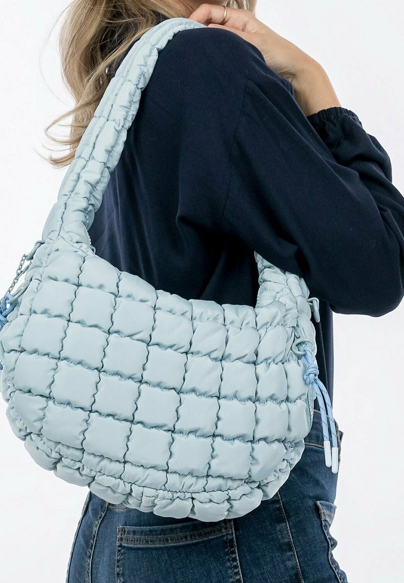 Femme portant un haut sombre et un jean porte un sac à bandoulière matelassé bleu clair avec une sangle épaisse.