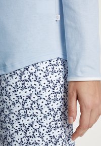 Lichtblauw langemouwen top met contrasterende witte afwerking, gecombineerd met een bloemenpatroon onderkant met marineblauwe bladeren op een lichtblauwe achtergrond.