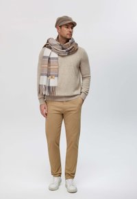 Maglione lavorato a maglia beige, chinos beige, sneakers bianche e una sciarpa decorata con righe color crema, grigio e marrone. Accessorio: berretto grigio.