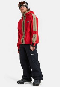 Man met een rode en beige snowboardjas, zwarte skibroek, zwarte beanie, skibril en zwarte sneeuwlaarzen, staande tegen een witte achtergrond.