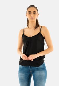 Top noir à bretelles spaghetti avec un décolleté en V, ornée de clous dorés le long du col. Le tissu est lisse et léger, se mariant parfaitement avec un jean bleu.