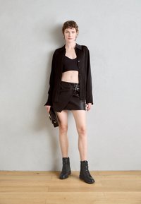 Chemise oversize en soie noire, top noir court, jupe en cuir noir ceinturée, bottines à lacets grises, tenant un petit sac à main noir.