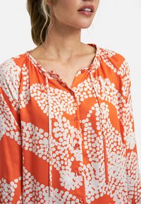 Oranžová blůza se vzorem bílých kruhů, s nařaseným výstřihem, zapínáním na knoflíky a dlouhými rukávy. Měkká látka s hladkým povrchem.