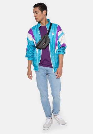 Veste coupe-vent teal avec des accents violets et blancs, t-shirt violet, jeans bleu clair, sac bandoulière noir et baskets blanches.