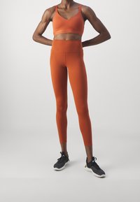Sostenitore sportivo arancione e leggings a vita alta abbinati realizzati in tessuto elasticizzato. I leggings sono aderenti e senza pattern visibili. Scarpe da atletica nere.