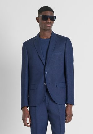 Completo blu navy realizzato in tessuto texturizzato, dotato di giacca monopetto con revers a trombetta e pantaloni abbinati. Occhiali da sole oversize neri.