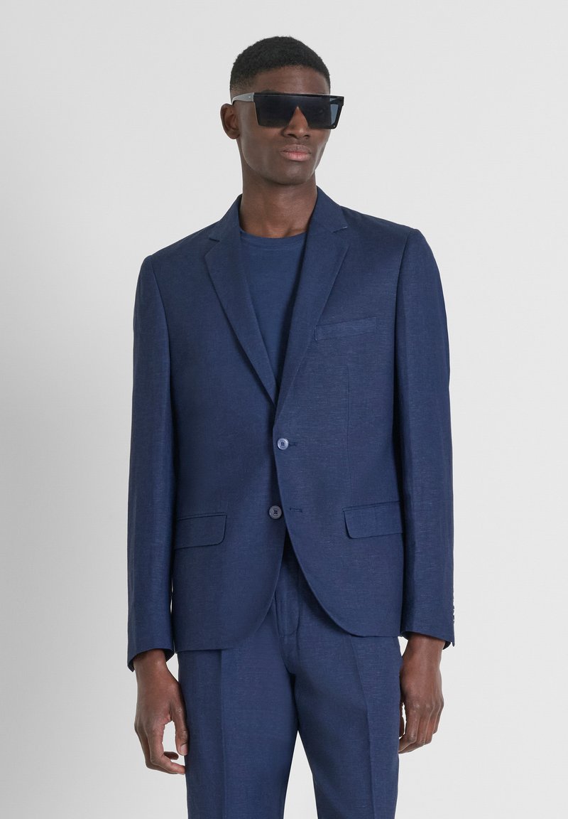 Traje azul marino hecho de tejido texturizado, que incluye una chaqueta de un botón con solapa muesca y pantalones a juego. Gafas de sol grandes y negras.