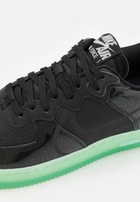 Svarta syntetiska sneakers med en glansig finish, utrustade med en grön genomskinlig sula, texturerad ovandel och framträdande logotyp på tungan.