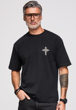 Homme aux cheveux gris et à la barbe, portant des lunettes noires, un t-shirt noir avec une croix ornée de bijoux, ainsi que des bracelets et une montre noirs, debout devant un fond uni.