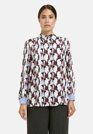 Woman wearing a long-sleeve button-up shirt with a multicolored abstract geometric pattern and pinstriped dark trousers against a white background.