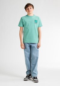 Mintgroen katoenen T-shirt met korte mouwen en een ronde hals, met een logo op de borst. Gecombineerd met lichtblauwe spijkerbroek en zwarte sneakers.