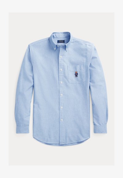 Chemise boutonnée bleu clair, en tissu coton, poche poitrine avec un design de ours brodé en bleu et noir, manches longues, ourlet arrondi.