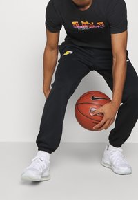 Camiseta negra con gráfico colorido, pantalones deportivos oscuros con el logo de los Lakers, zapatillas blancas deportivas, sosteniendo un balón de baloncesto en posición agachada.