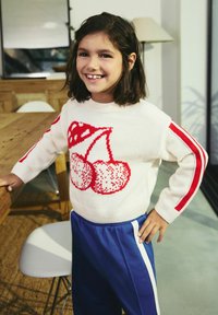 Pull en laine crème avec motif de cerises rouges et manches rayées rouges, associé à un pantalon de survêtement bleu avec des rayures blanches sur les côtés.