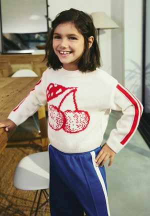 Maglione lavorato a maglia color crema con stampa di ciliegie rosse e maniche a strisce rosse, abbinato a pantaloni da jogging blu con strisce bianche sui lati.