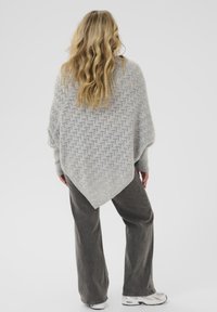 Hellgrauer gestrickter Poncho mit strukturiertem Muster, mit spitzem Saum und oversized Passform, kombiniert mit dunklen Schlaghosen und Sneakers.