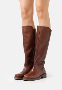 Botas de cuero marrón hasta la rodilla con una textura suave, puntera redondeada, cremallera lateral y un tacón bajo en bloque.