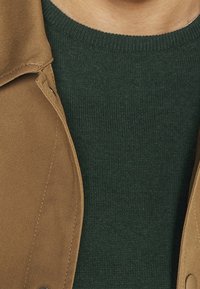 Beige tygkappa med krage, över en mörkgrön ribbad stickad tröja. Har synlig sömnad och en knapp på vänster sida av kappan.