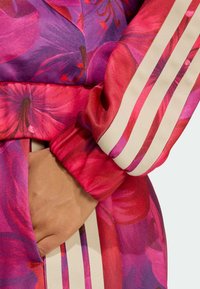 Veste de sport fleurie dans des tons de rose et de violet, avec des poignets rayés et une texture douce, une main placée dans une poche latérale.