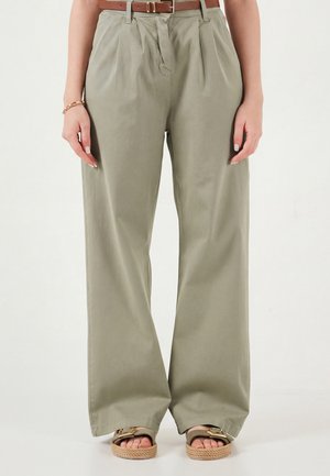 Femeie în picioare purtând pantaloni khaki cu talie înaltă și talie largă, cu curea maro și sandale cu tălpi din sfoară, pe un fundal simplu.