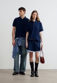 Νεαρός άνδρας και γυναίκα που φορούν μπλε μπλουζάκια πόλο, η γυναίκα με πλισέ φούστα και μπότες κρατάει μια κόκκινη χειραποσκευή, ο άνδρας με πουκάμισο δεμένο γύρω από τη μέση.