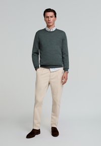 Grijze gebreide trui over een witte blouse, beige broek en donkerbruine schoenen. Zachte textuur en casual pasvorm door het geheel.