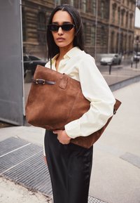 Borsa tote in suede marrone con una forma strutturata, dotata di una tracolla in pelle scura e dettaglio con fibbia. Portata da una persona che indossa una camicia bianca.