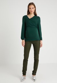 Top de maternidade verde com design em cruz e mangas longas, combinado com calças justas verde-escuras e sapatos de salto agudo prateados.