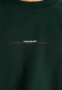 Sudadera de un verde oscuro con texto blanco parcialmente cubierto por una línea horizontal morada cerca del área del pecho.