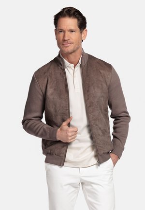 Man wearing a brown zip-up jacket over a white polo shirt with white pants, standing with one hand in pocket and the other adjusting jacket.