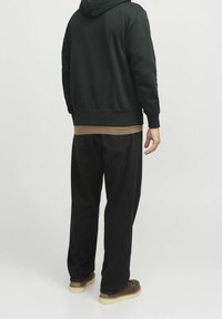 Ciemnozielona bluza z długim rękawem z prążkowanymi mankietami, nałożona na beżową koszulę, w połączeniu z luźnymi czarnymi spodniami i brązowym obuwiem.